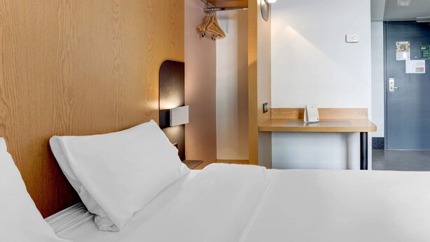 Ein Hotelzimmer mit einem ordentlich bezogenen Bett, einem Holz-Kopfteil, einem Nachttisch mit Lampe und einer Tür mit Griff.