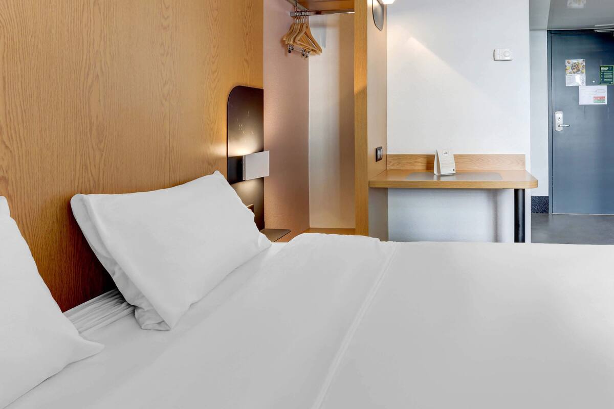 Ein Hotelzimmer mit einem ordentlich bezogenen Bett, einem Holz-Kopfteil, einem Nachttisch mit Lampe und einer Tür mit Griff.