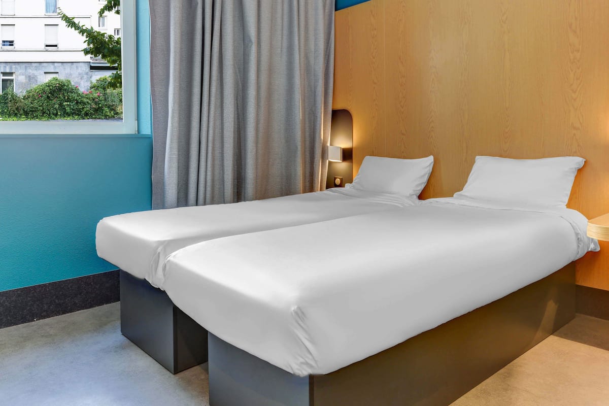 Ein Hotelzimmer mit zwei Einzelbetten, einem Fenster mit Vorhängen, einem Nachttisch mit Lampe und Blick auf Gebäude und Grünflächen.