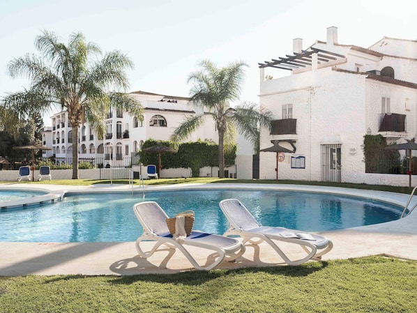 A swimming pool with lounge chairs, palm trees, and a building with balconies. - Apartamentos Ona Club Bena Vista (Estepona)