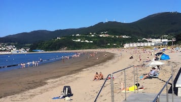 Una playa con una escalera que baja a la arena, cielo despejado y un paisaje montañoso al fondo.