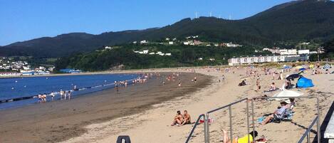 A beach with a staircase leading down to the sand, a clear sky, and a hilly landscape in the background.