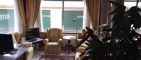 A room with a TV, a striped armchair, a small table, a potted plant, and a large window with curtains.