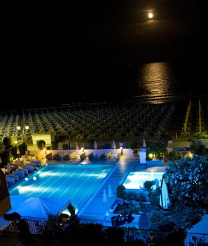 A nighttime view of a pool area with lounge chairs and umbrellas, surrounded by a fence and trees. - Hotel Sirenetta (Jesolo)
