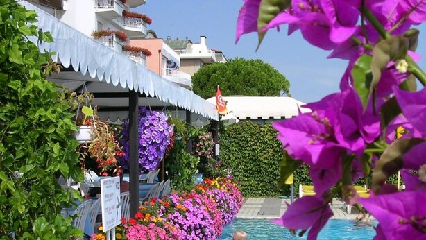 A swimming pool surrounded by vibrant flowers and a white building with balconies. - Hotel Sirenetta (Jesolo)