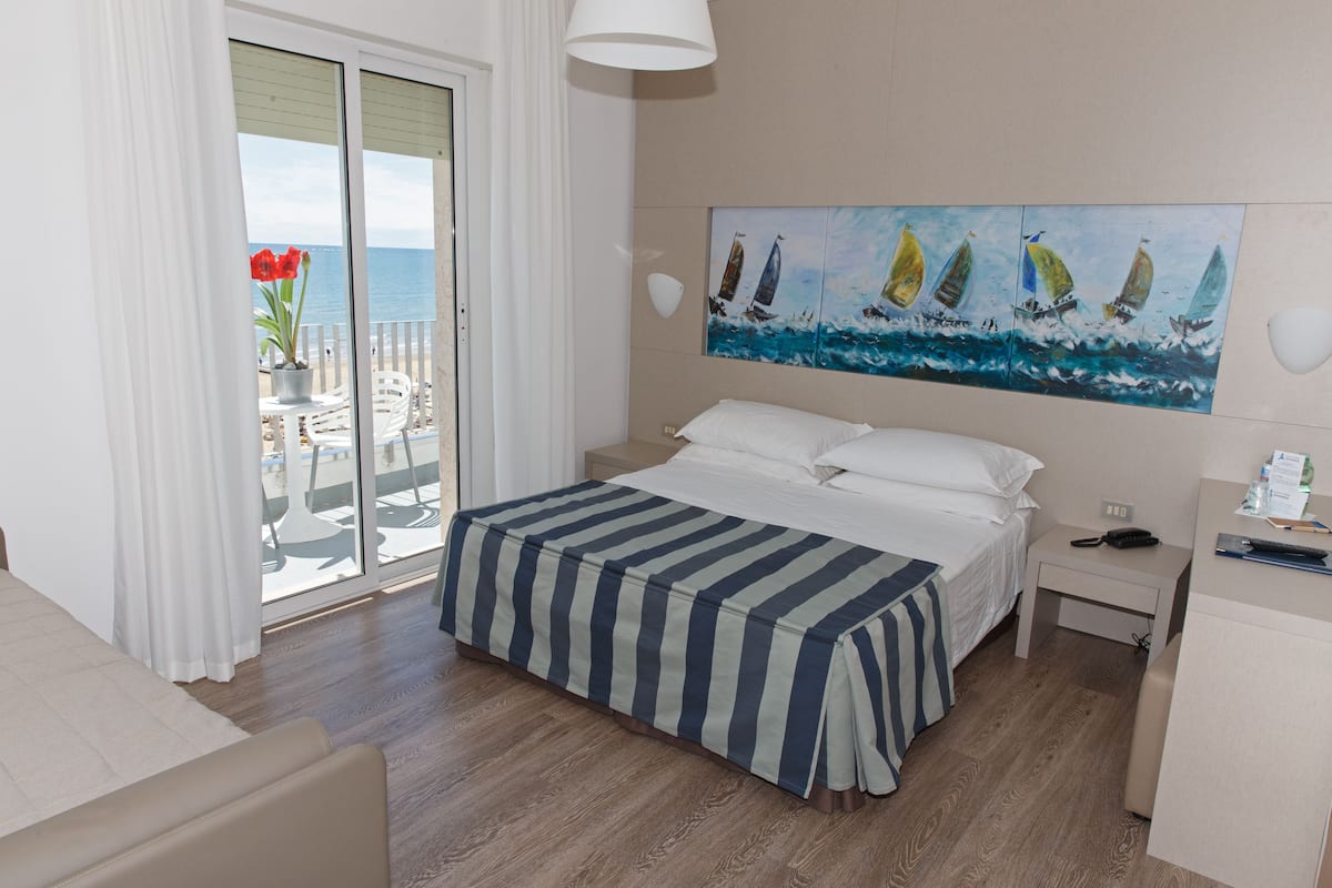 A hotel room with a bed, a desk, a chair, a painting of sailboats, and a view of the sea through a sliding door.