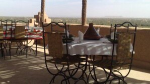 An outdoor dining area with a table set for two, surrounded by wicker chairs, under a woven roof, overlooking a distant landscape. - Dar Panorama (Skoura)