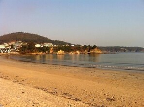 A sandy beach with a calm sea, a few scattered rocks, and a distant hill with buildings. - Hotel As Areas II (Viveiro)