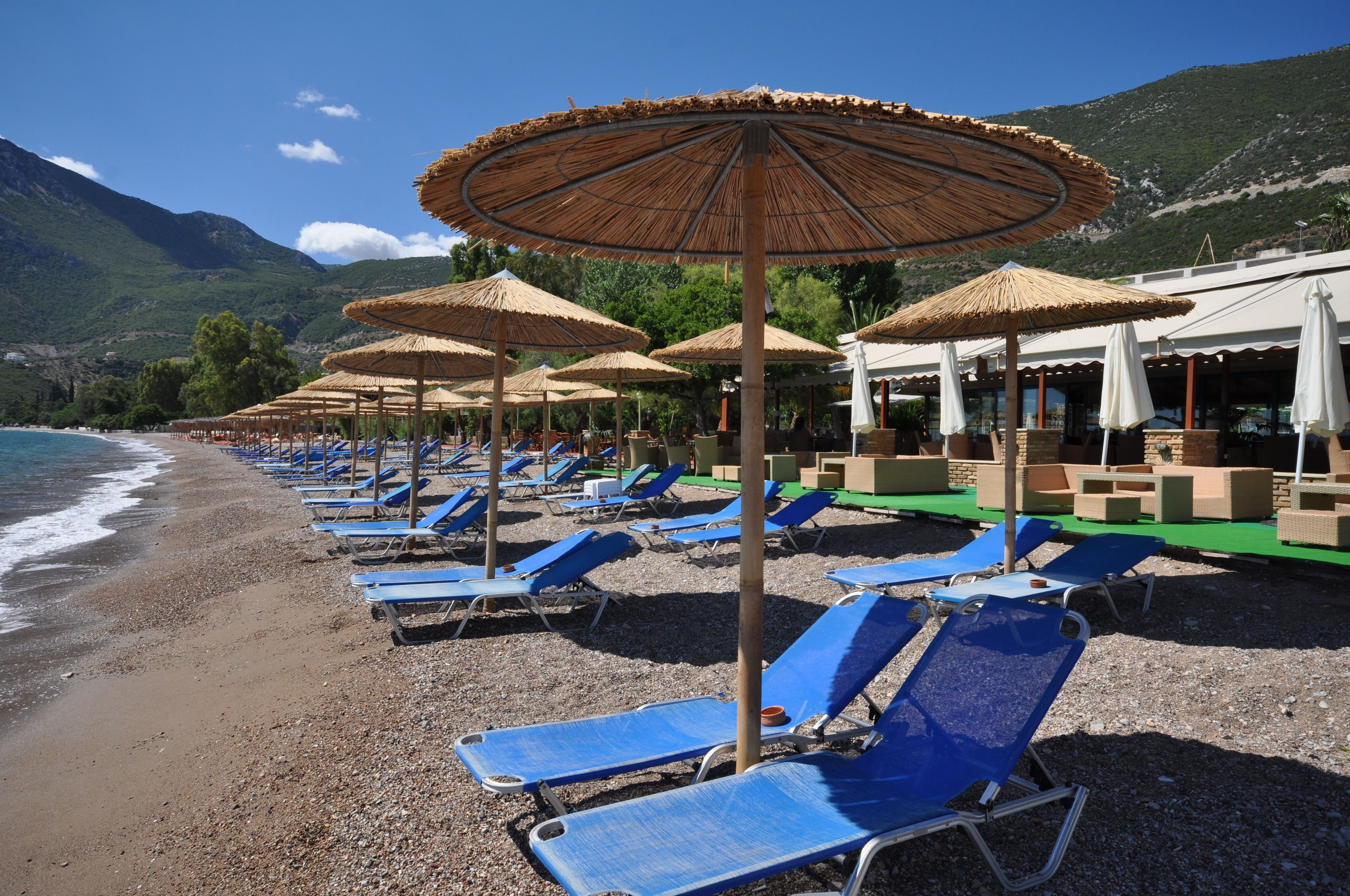 A beach with rows of blue sun loungers and thatched umbrellas, a pebble beach, and a mountain in the background.