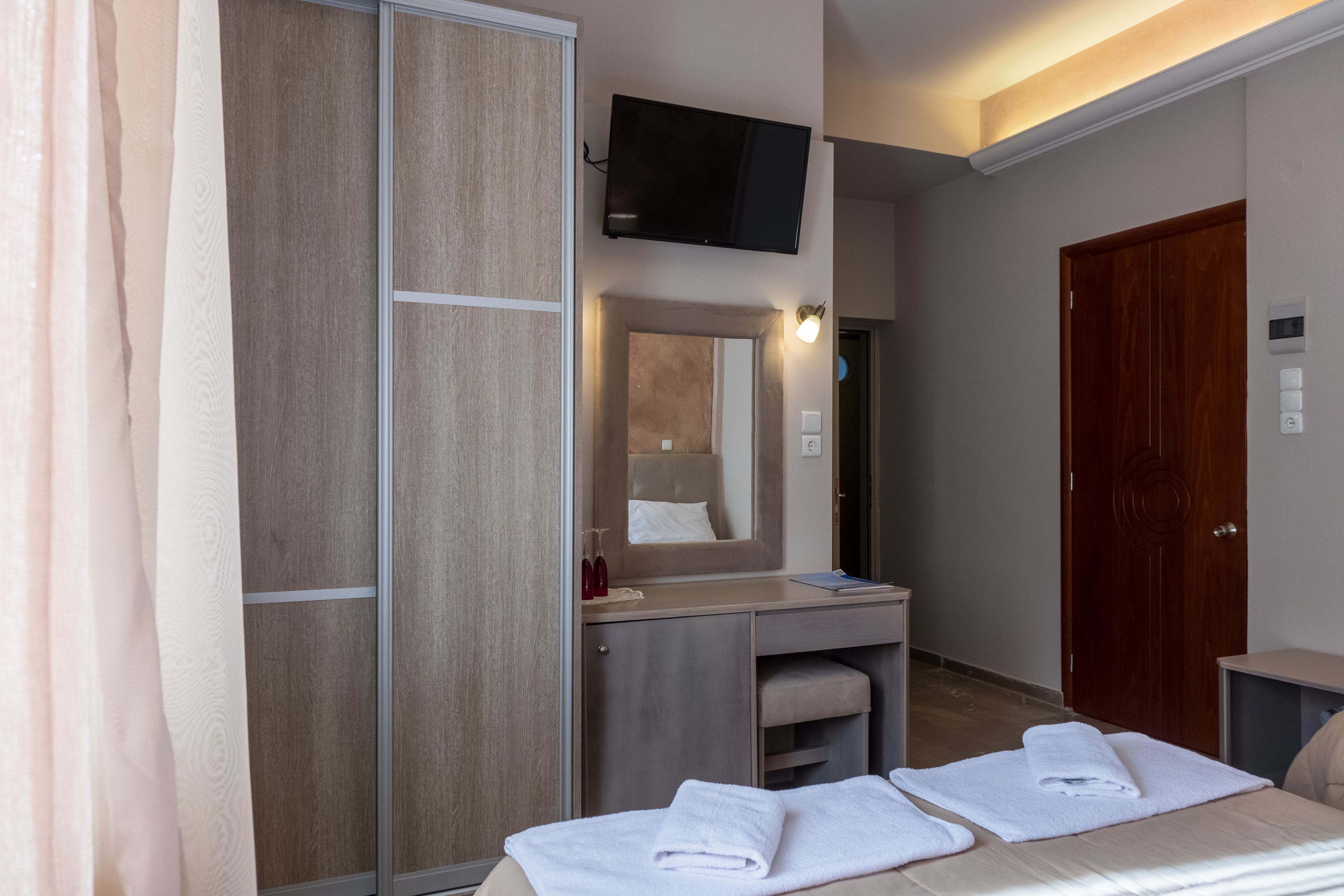 A hotel room with a bed, a TV mounted on the wall, a wooden wardrobe, a mirror, and a door.