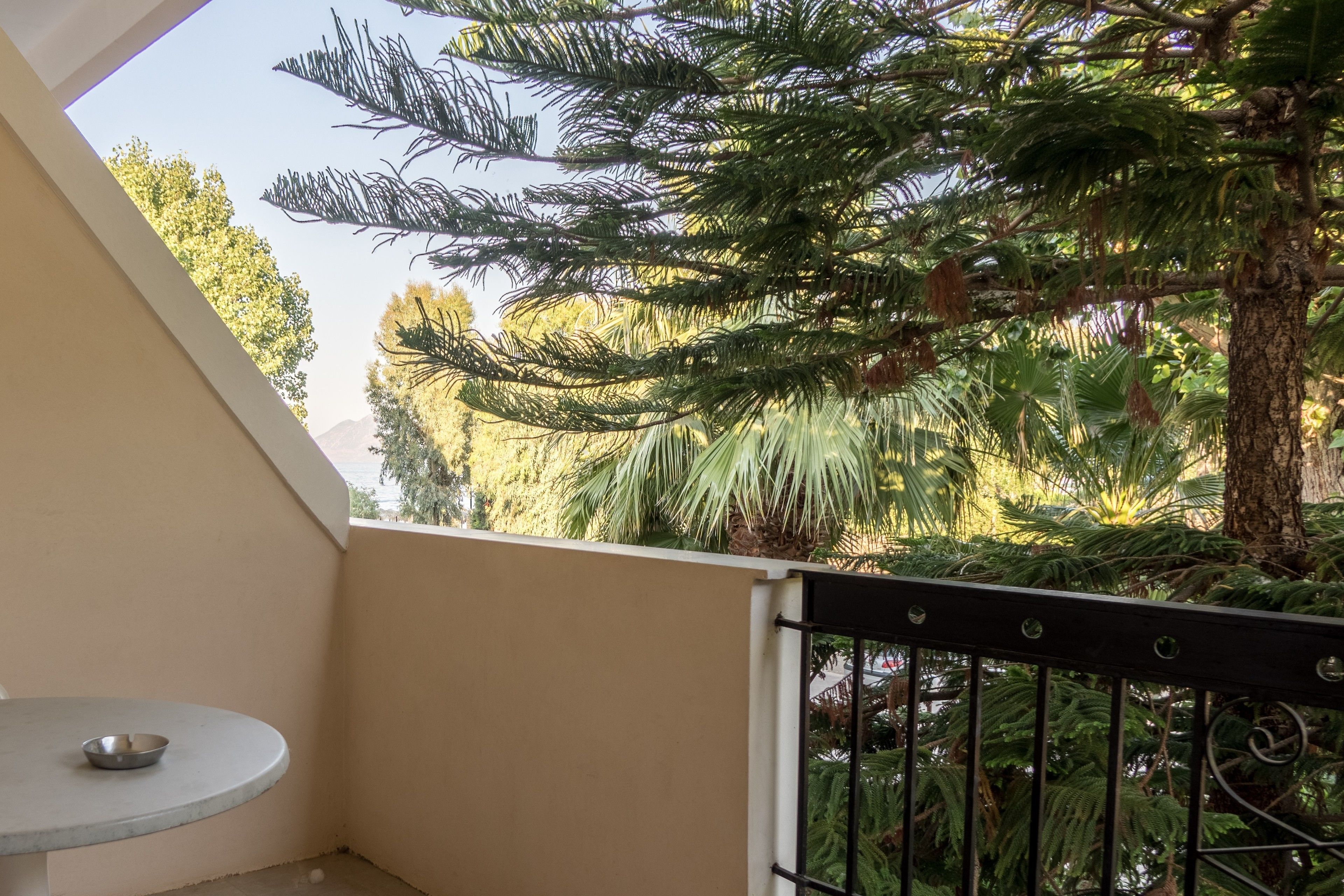 A balcony with a round table and a metal railing, overlooking a lush garden with various trees and plants.