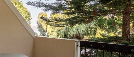 A balcony with a round table and a metal railing, overlooking a lush garden with various trees and plants.
