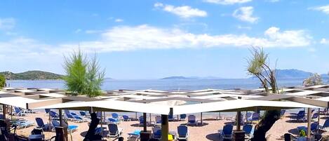 Een zitgedeelte aan het strand met parasols en ligbedden, met uitzicht op een waterpartij en verre bergen.