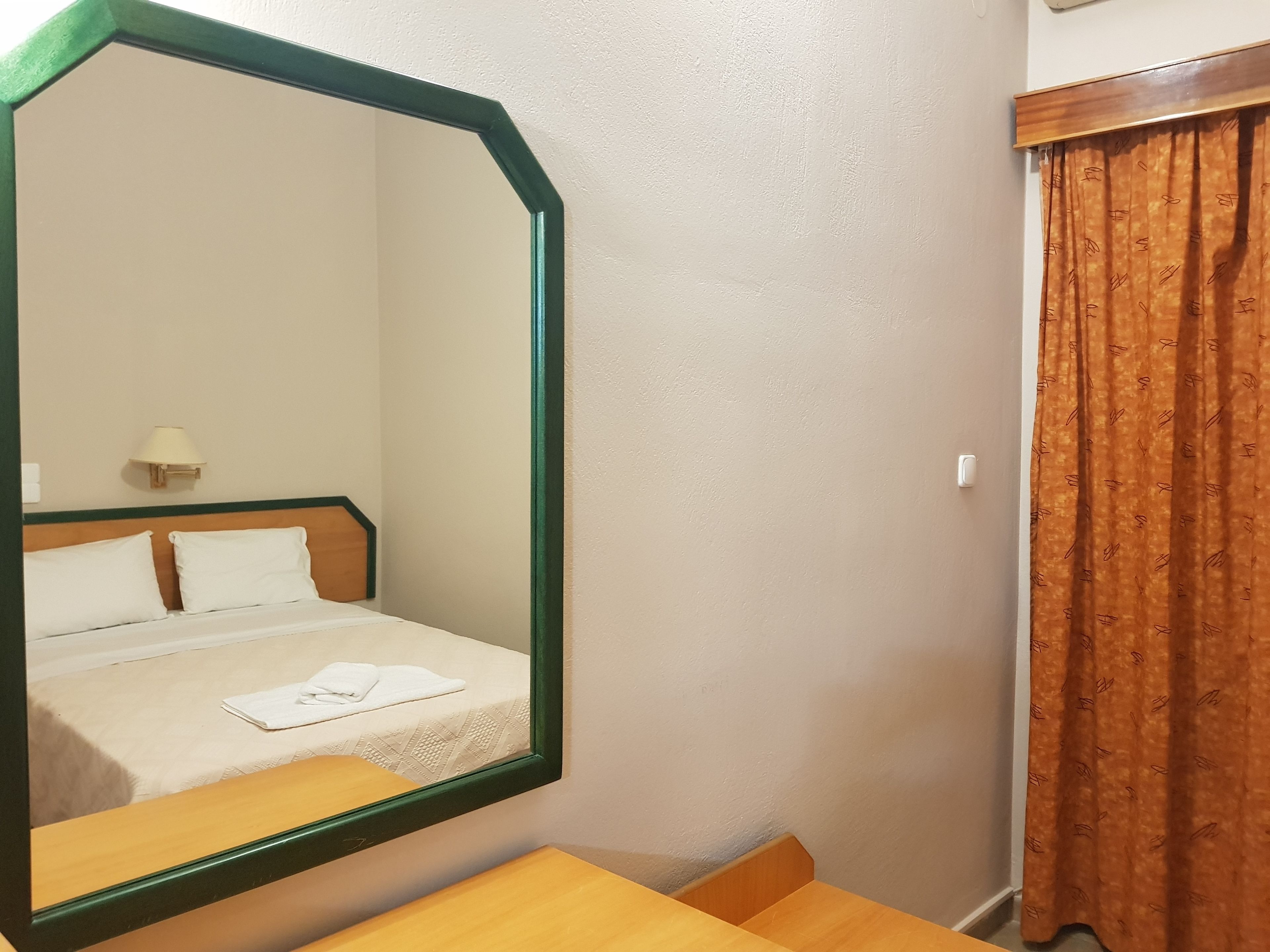Habitación de hotel con una cama, una mesita de noche de madera, un espejo, una lámpara de pared y una ventana con cortinas.