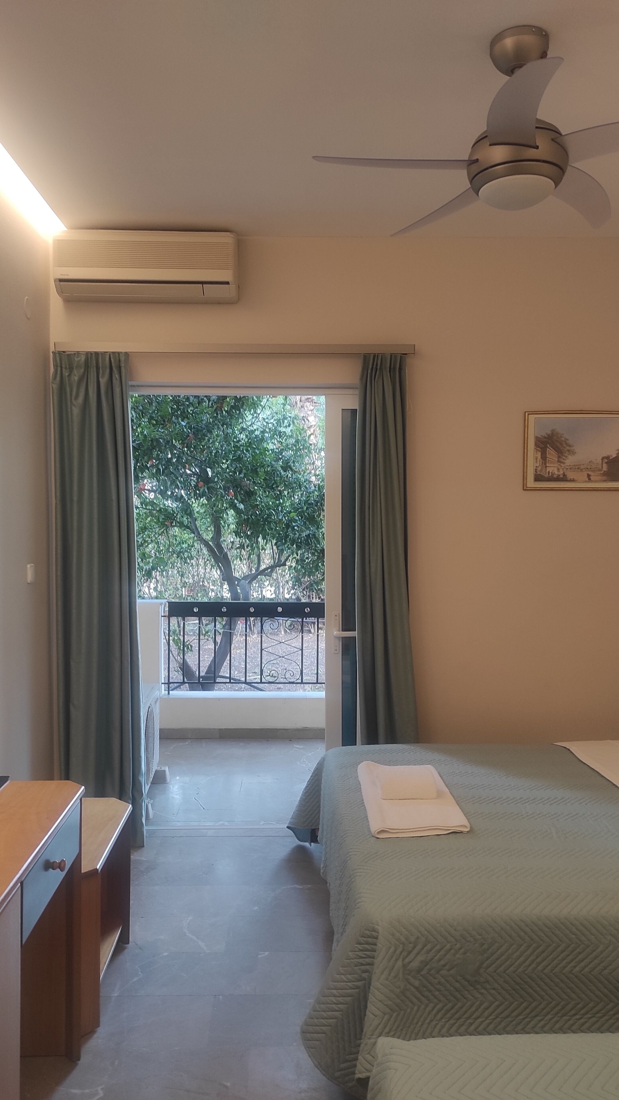 A hotel room with a bed, a desk, an air conditioner, a ceiling fan, and a balcony with a view of trees.