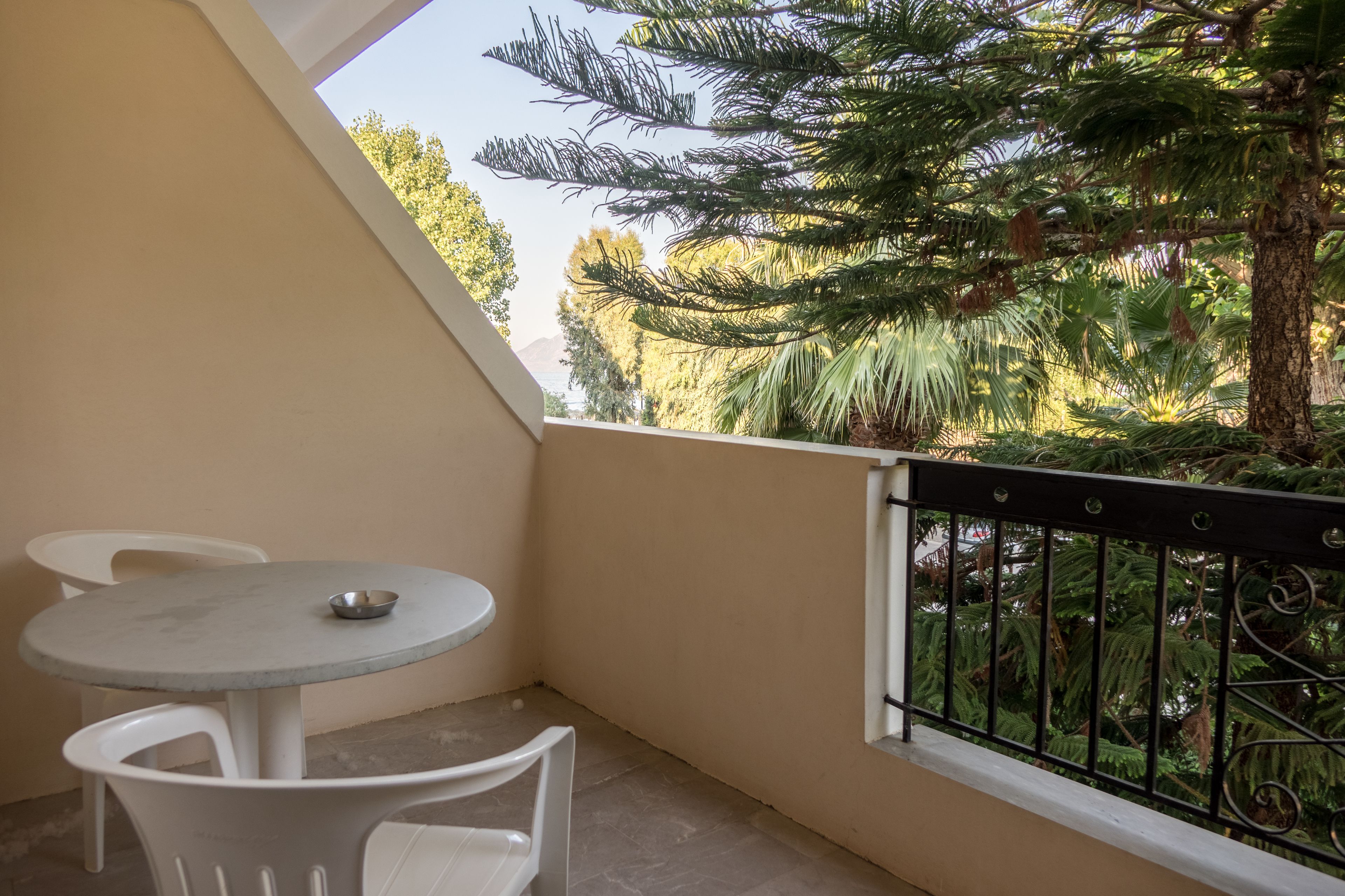 Een balkon met een ronde tafel en twee stoelen, een metalen hekwerk, en uitzicht op een dennenboom en andere begroeiing.
