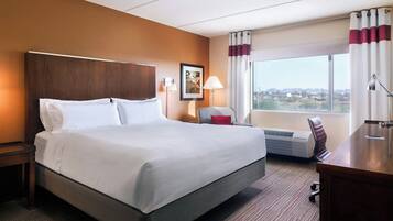 Habitación de hotel con una cama grande, un escritorio, una silla, una lámpara, un cuadro en la pared y una ventana con vistas.