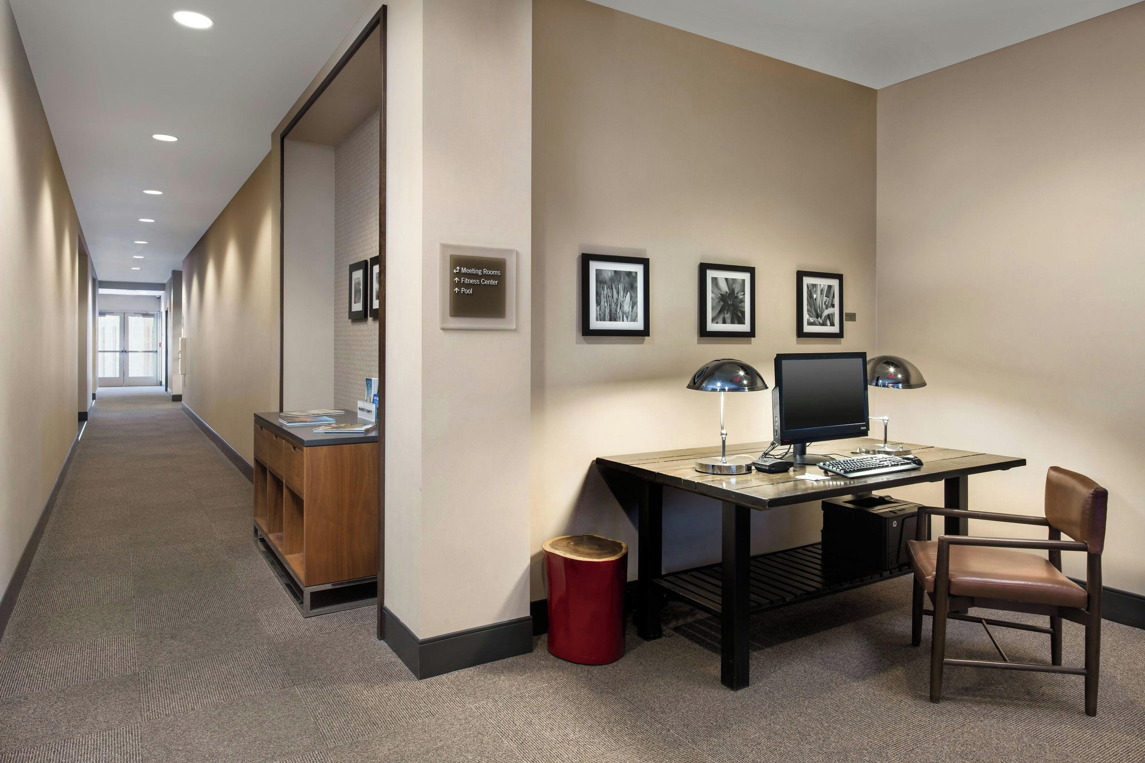 A hotel corridor with a desk, chair, and computer setup. There are framed pictures on the wall and a sign indicating amenities like meeting rooms, fitness center, and pool.
