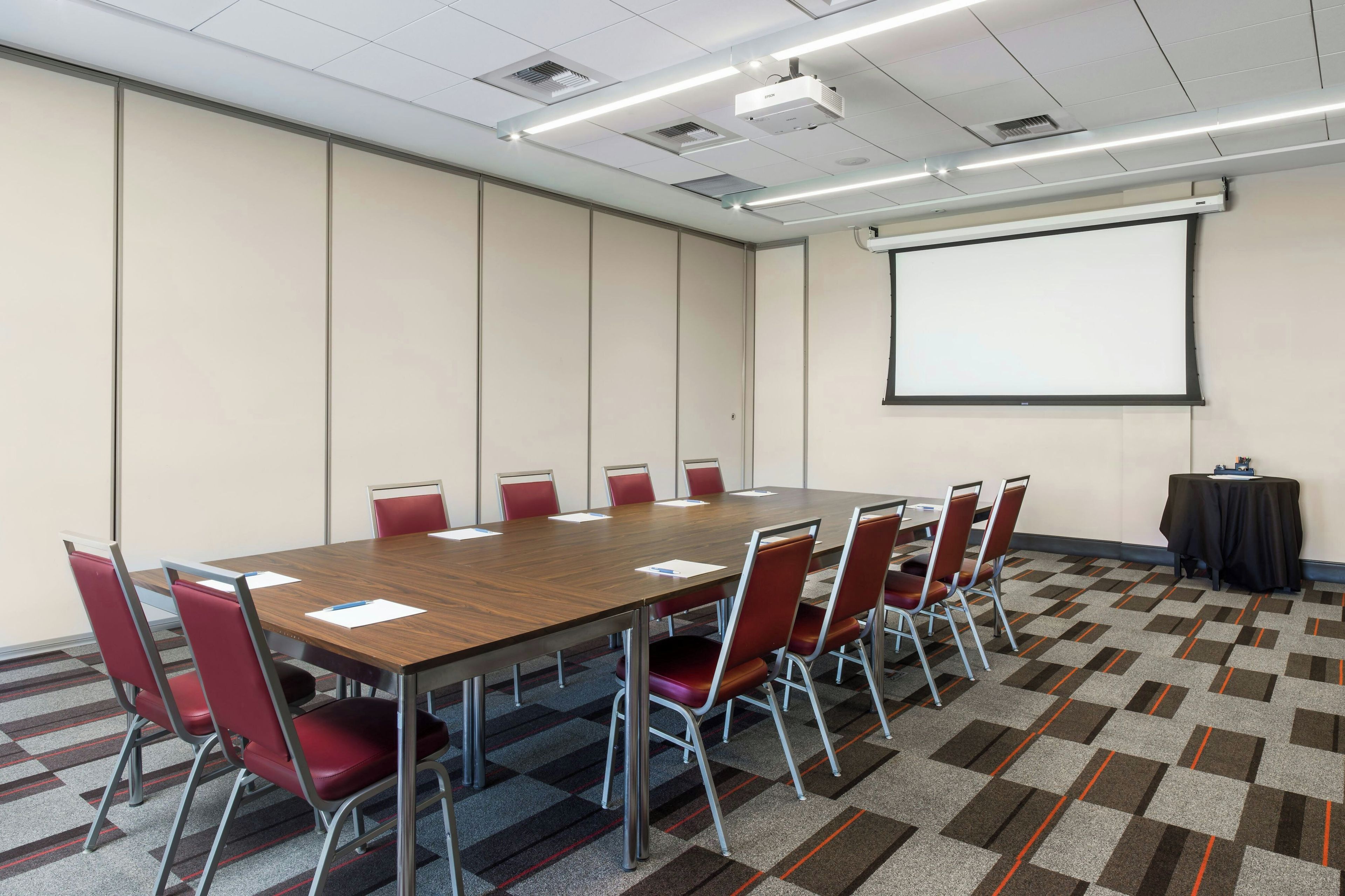 A conference room with a long table, red chairs, a projector screen, and a carpeted floor.
