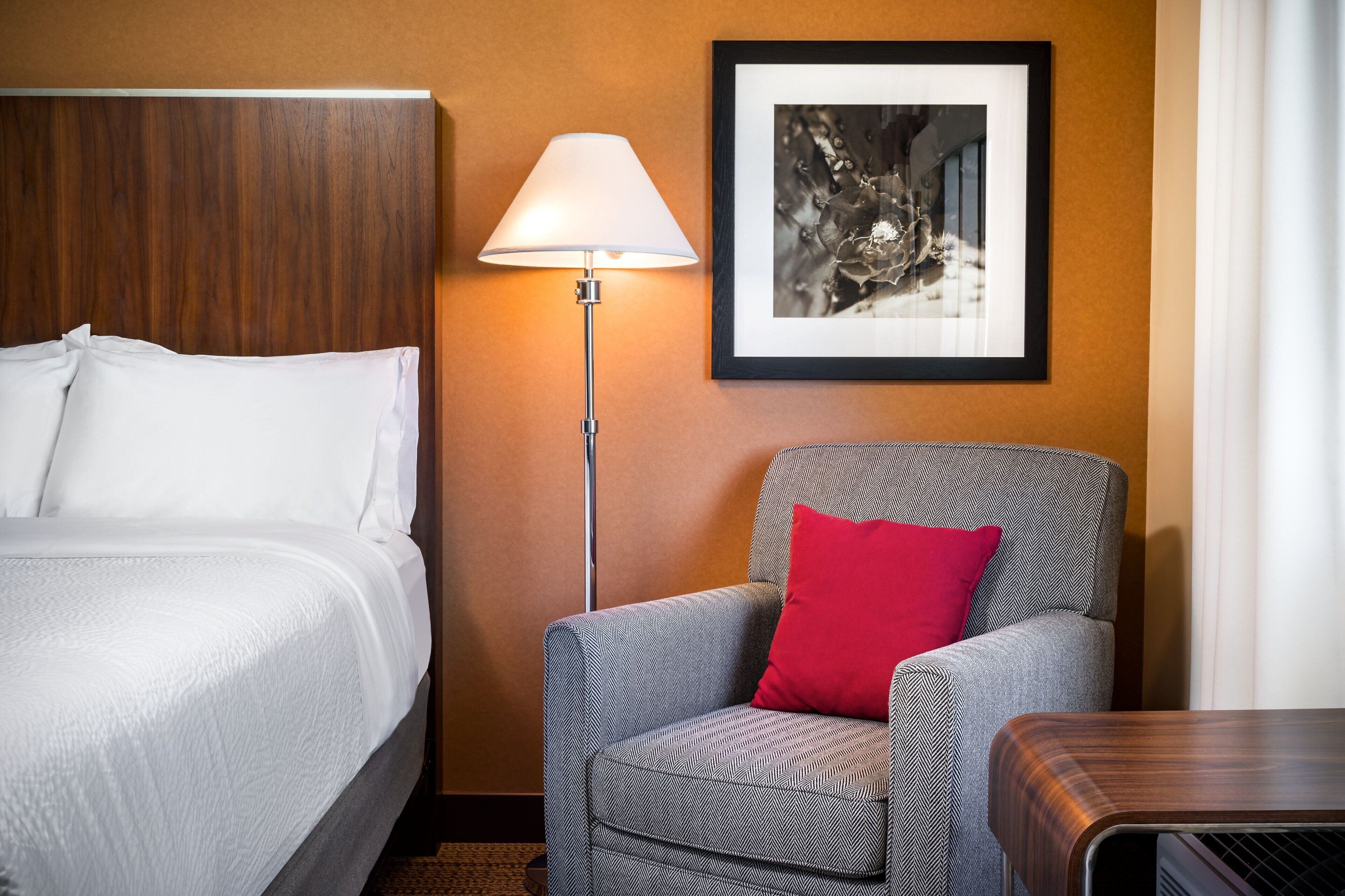 A hotel room with a bed, a chair with a patterned cushion, a standing lamp, and a framed picture on the wall.