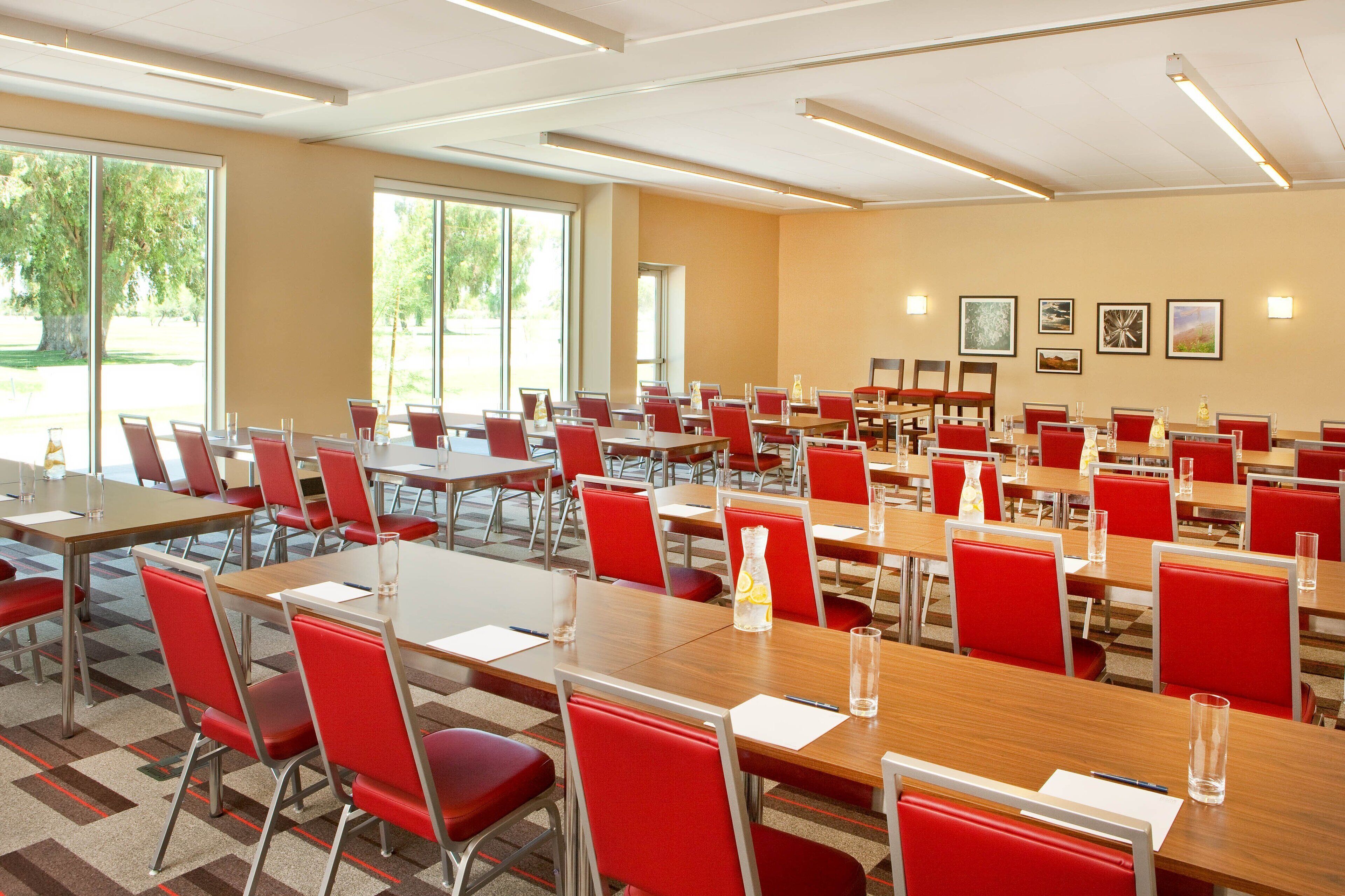A conference room with multiple tables and red chairs, large windows, and framed pictures on the walls.