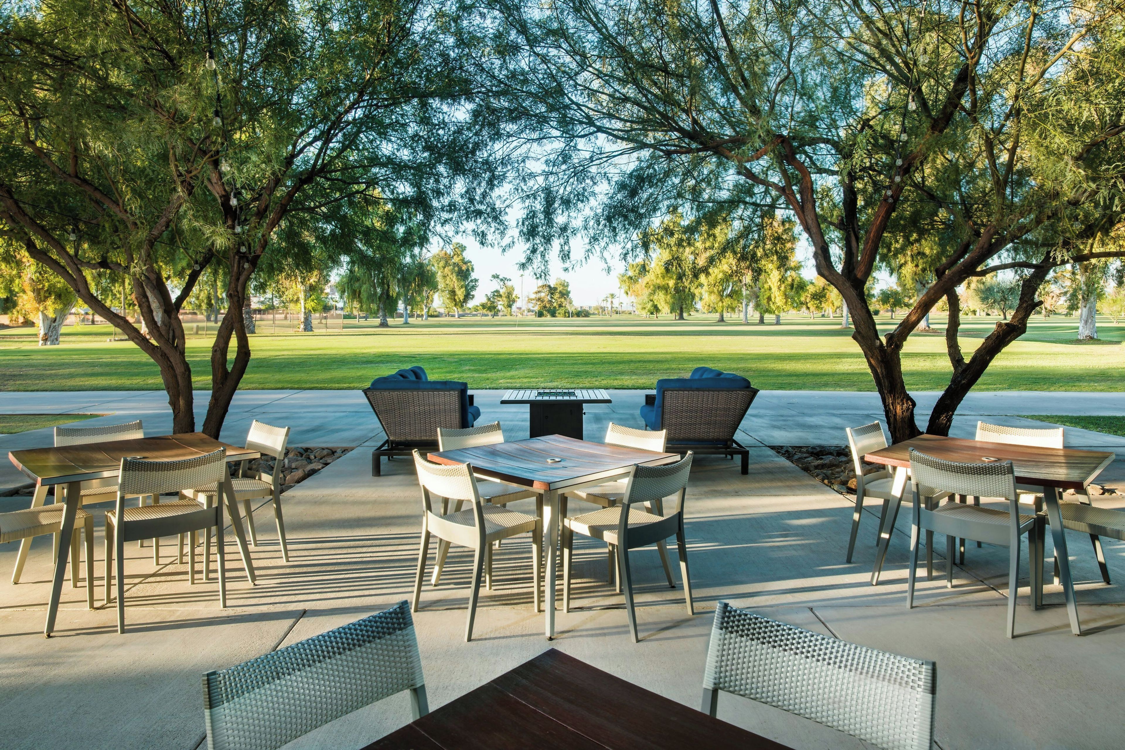 An outdoor patio area with tables and chairs, surrounded by trees, overlooking a grassy field.
