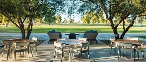 An outdoor patio area with tables and chairs, surrounded by trees, overlooking a grassy field.