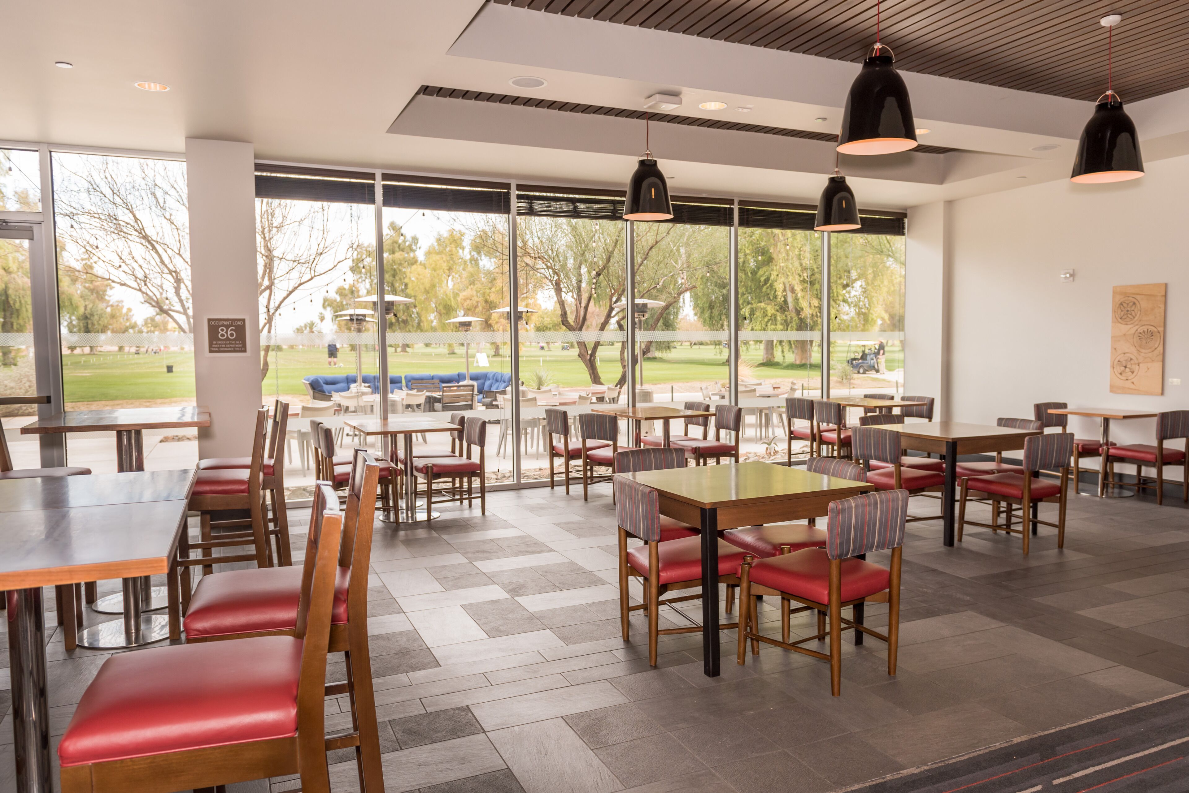 A spacious indoor dining area with multiple tables and chairs, large windows offering a view of the outdoors, and pendant lighting.