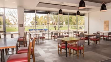 A spacious indoor dining area with multiple tables and chairs, large windows offering a view of the outdoors, and pendant lighting.