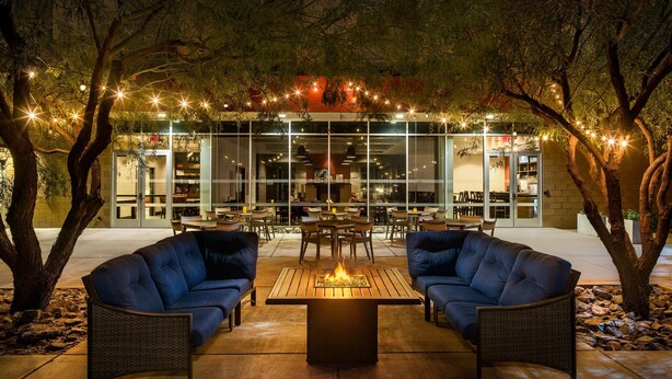 An outdoor seating area with a fire feature, surrounded by trees and a building with large windows and string lights.