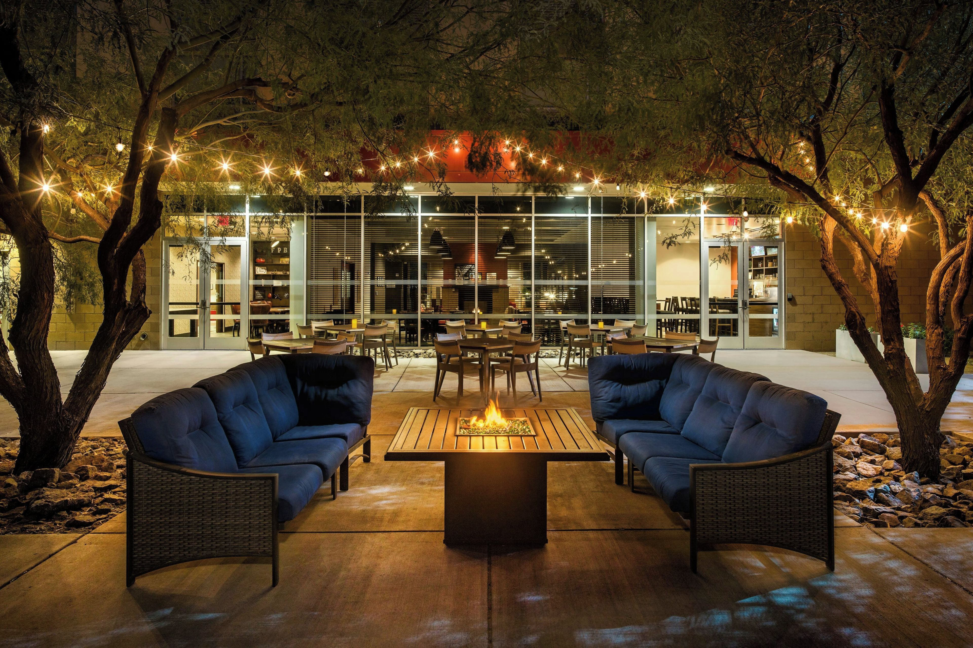 An outdoor seating area with two sofas and a central table with a fire feature, surrounded by trees and string lights.