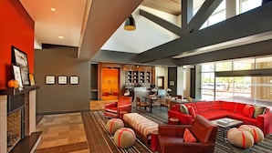 A modern hotel lobby with a fireplace, red seating, and a kitchen area visible through an open door.