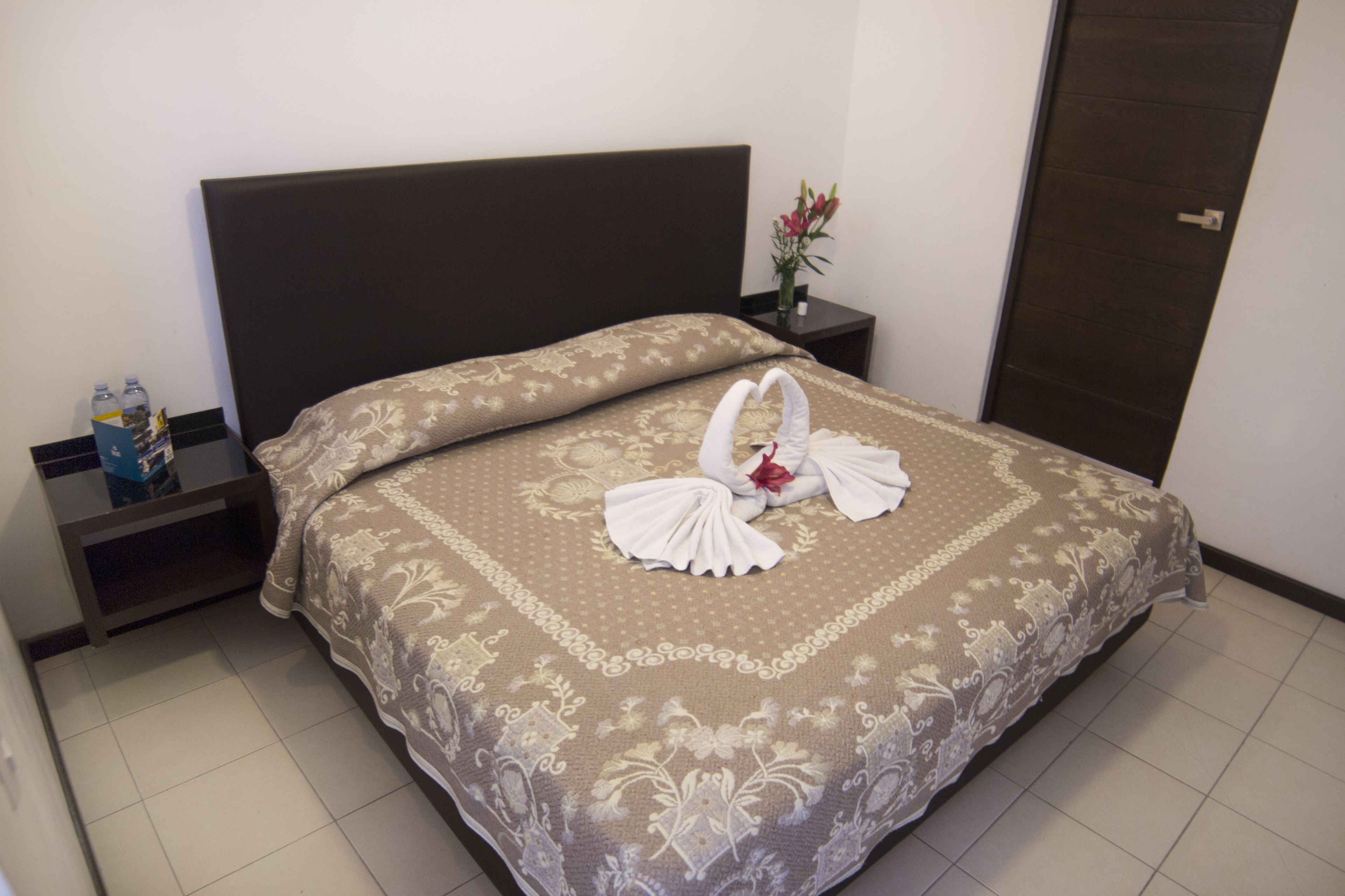Una cama bien hecha con una colcha estampada con motivos florales, una mesita de noche con un florero y una botella de agua.