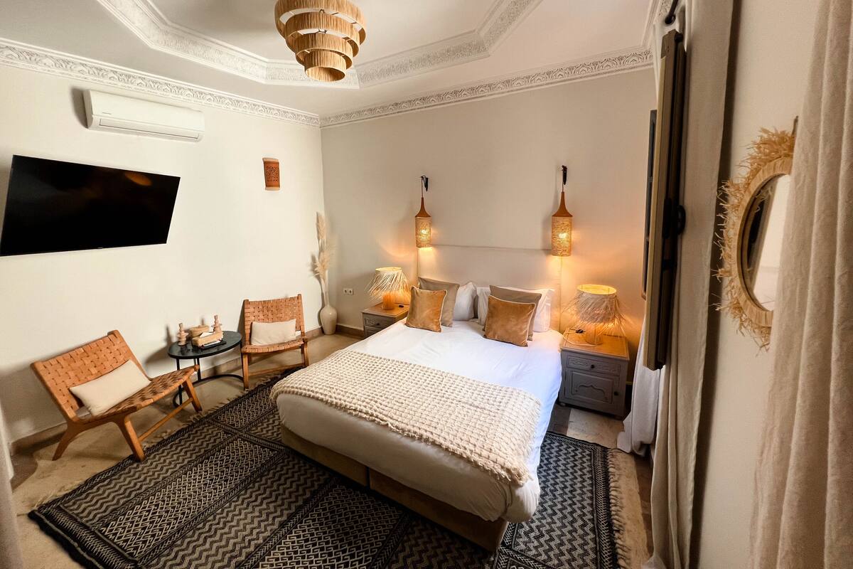 A bedroom with a large bed, two bedside tables, a TV mounted on the wall, and a ceiling light fixture.
