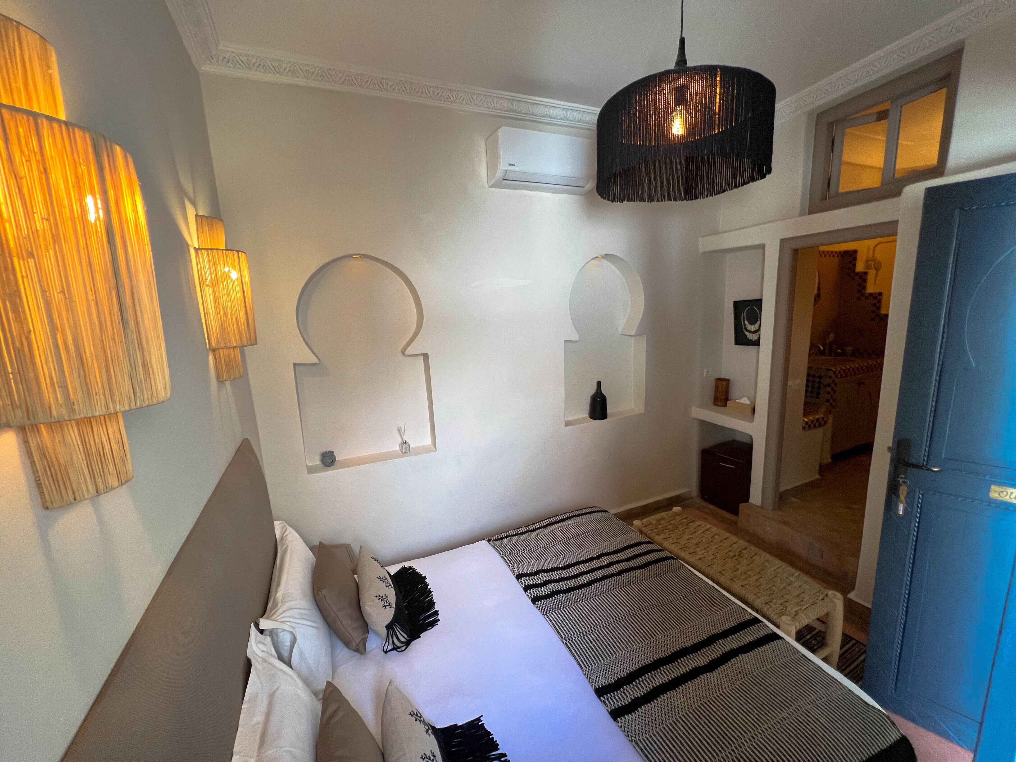 A bedroom with a bed, bedside lamps, a hanging light fixture, a mirror, and a door leading to another room.