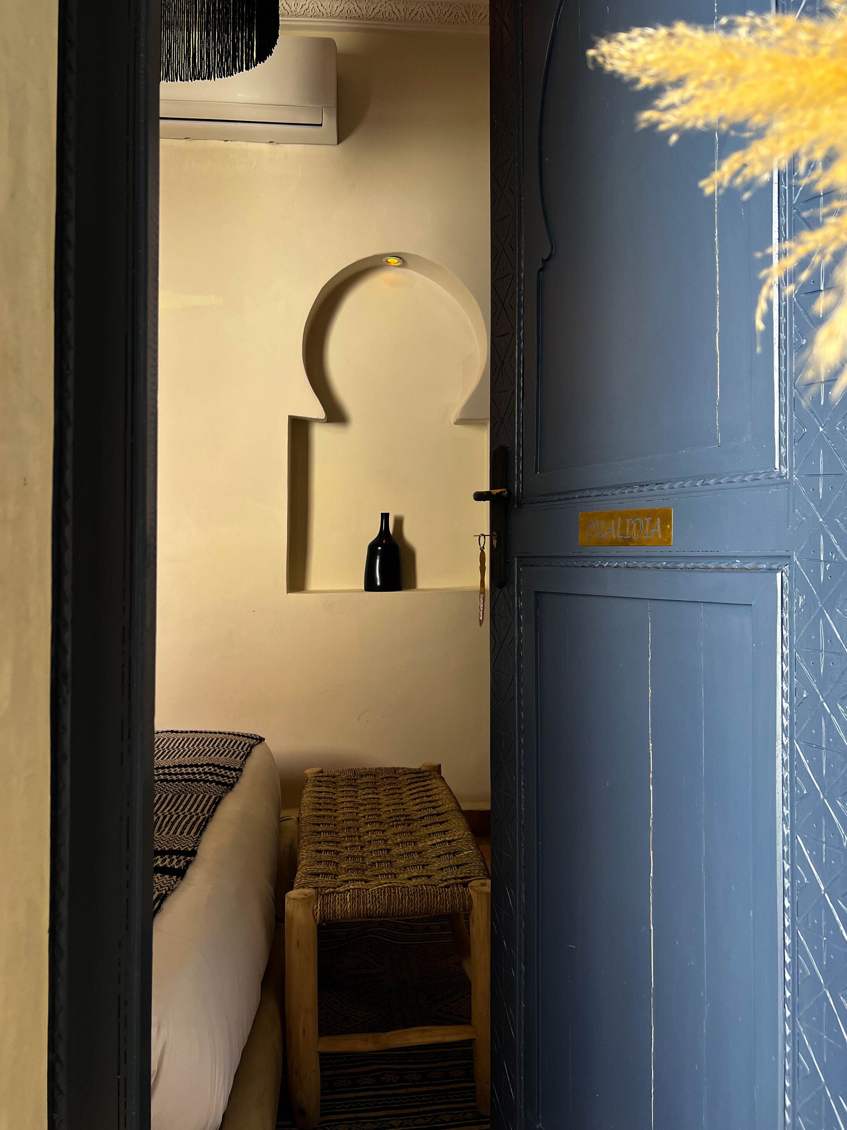 Un estrecho pasillo que da a una habitación con una cama, un taburete pequeño, un estante con una botella y una puerta con un cartel.