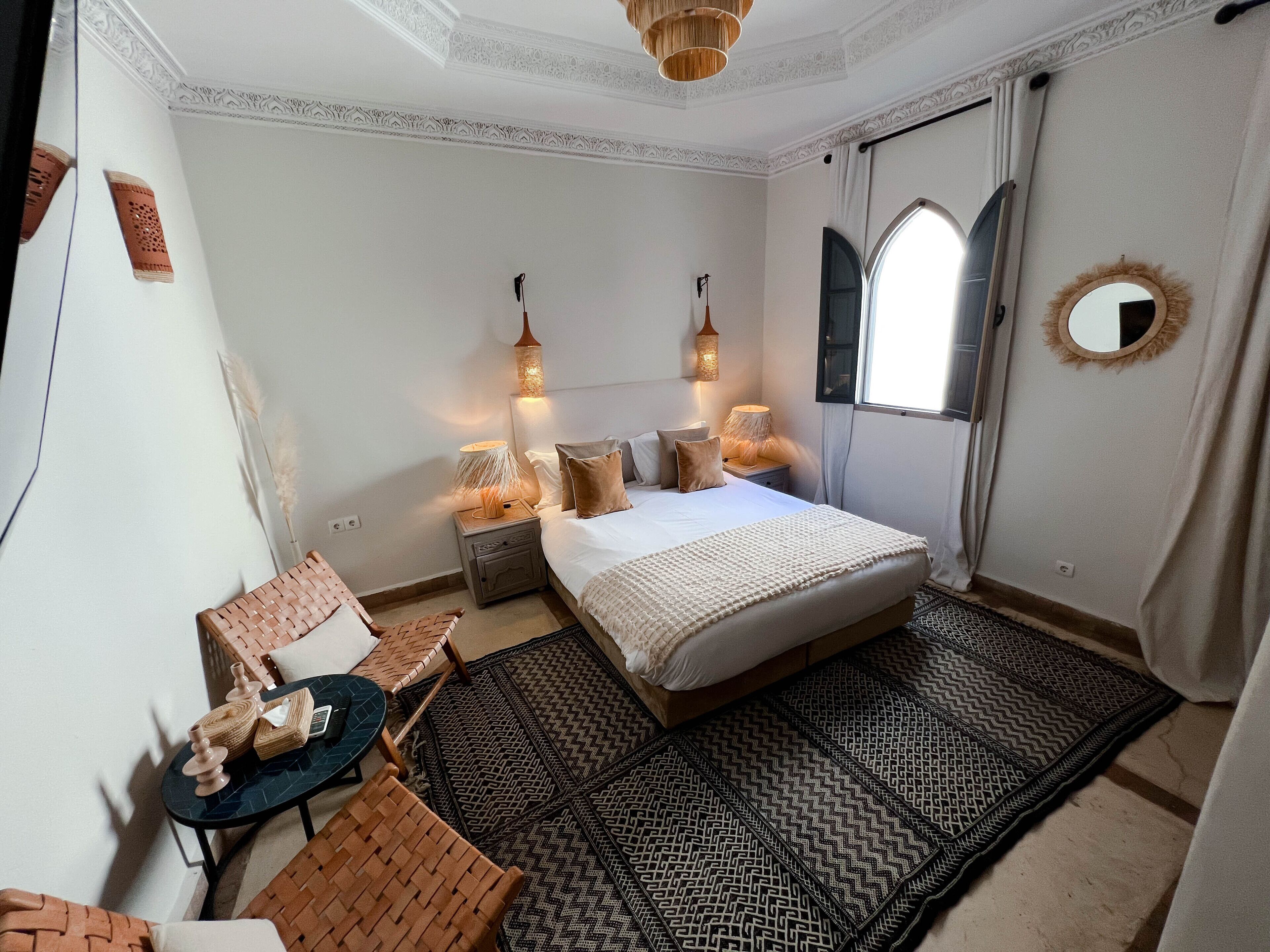 Una camera da letto con un letto grande, due comodini con lampade, un tavolino rotondo con sedie, un tappeto decorativo e una finestra con tende.