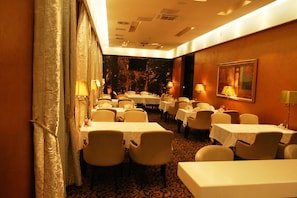 A hotel dining area with multiple tables and chairs, a patterned carpet, and wall-mounted lamps.