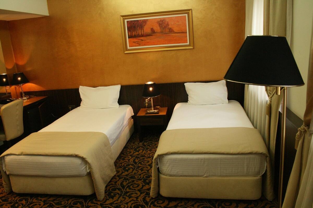 A hotel room with two single beds, a nightstand with lamps, a painting on the wall, and a patterned carpet.