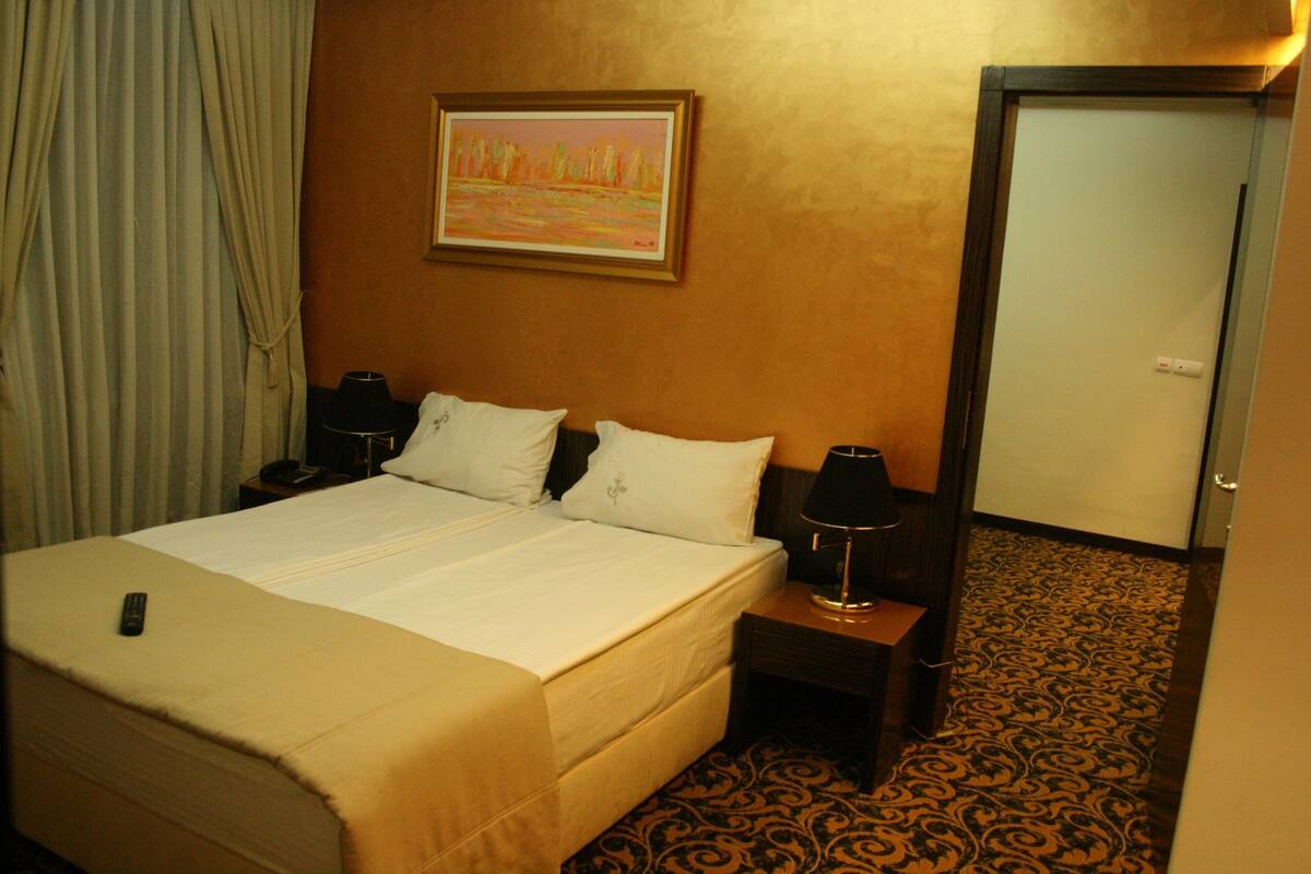 A hotel room with a bed, two bedside tables, a nightstand, a lamp, a painting on the wall, and a door.