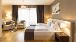 A hotel room with a bed, two chairs, a small table with a lamp, a bedside table with a lamp, a window with curtains, and a painting on the wall. - Empress Hotel (Munich)