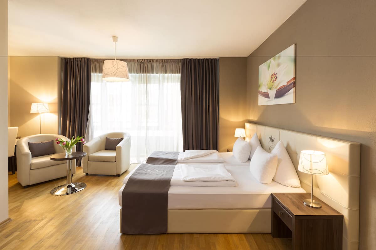 A hotel room with a bed, two chairs, a small table with a lamp, a bedside table with a lamp, a window with curtains, and a painting on the wall.