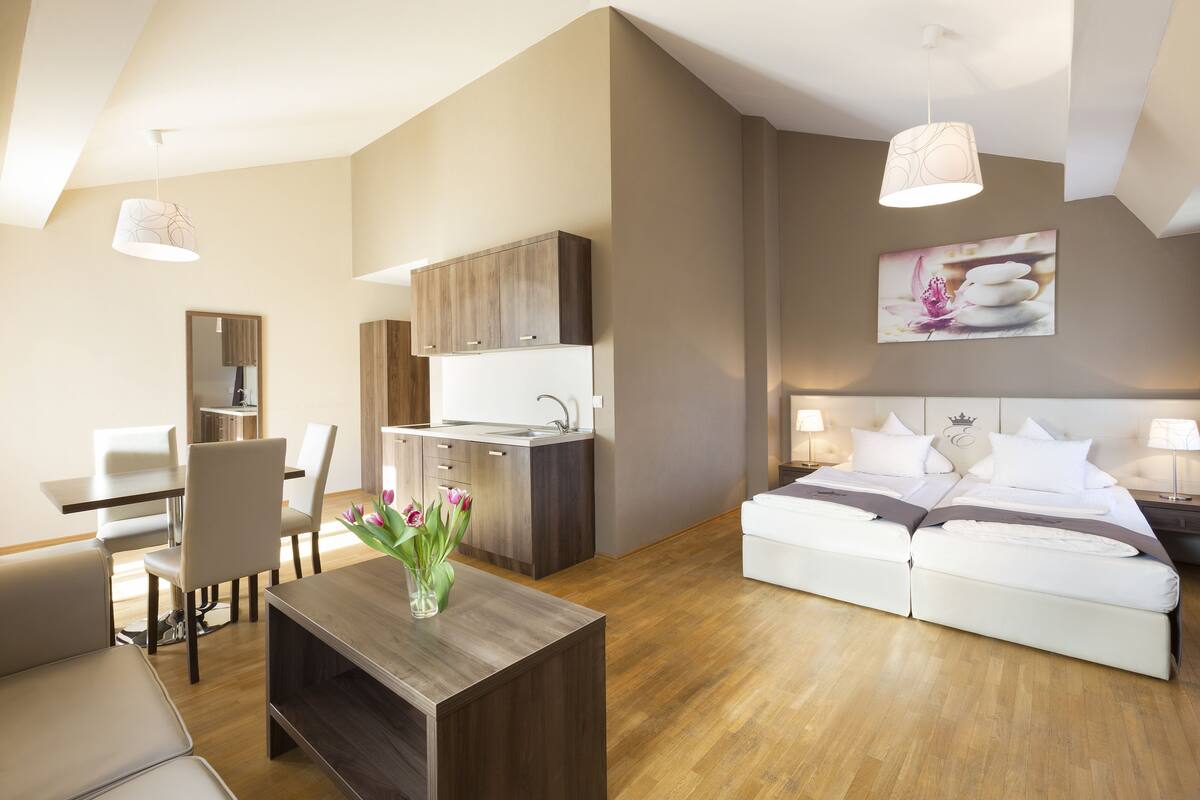 A modern hotel room with a dining area, kitchenette, and a large bed with white bedding.