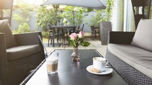 An outdoor seating area with wicker furniture, a table with a drink and a vase of flowers, and a patio umbrella. - Empress Hotel (Munich)