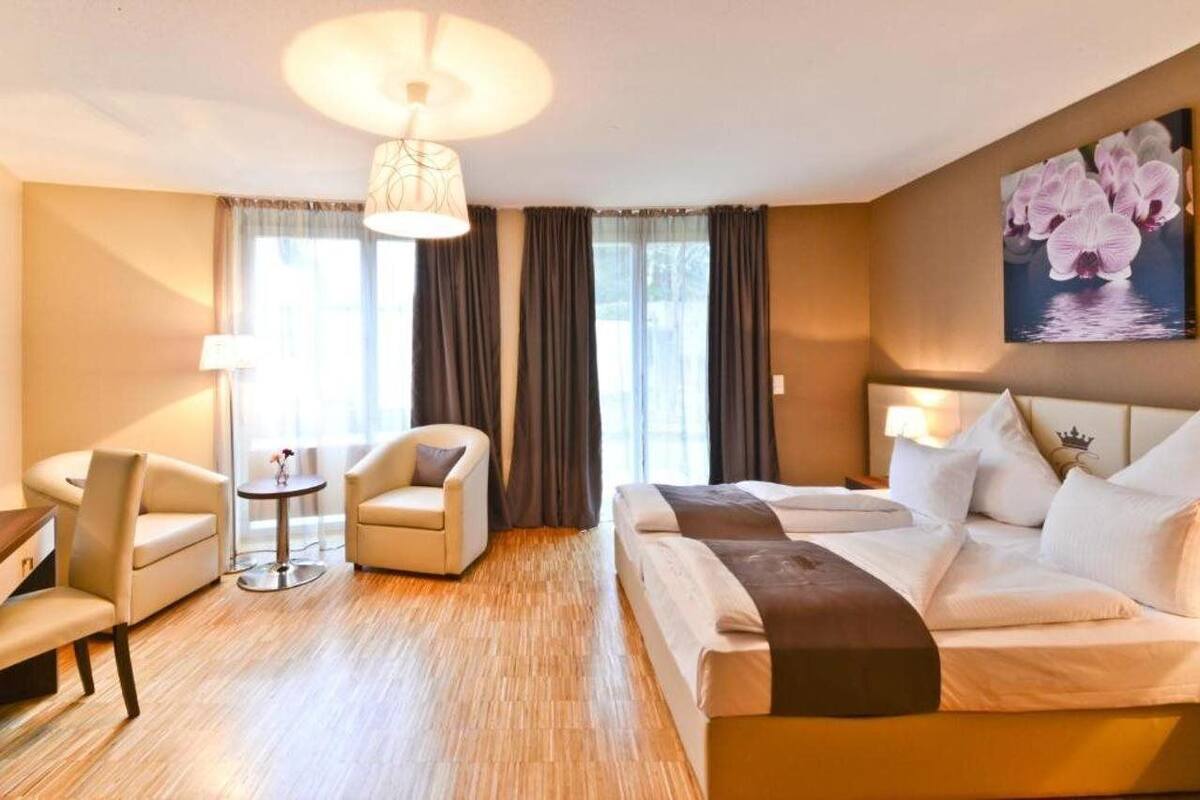 A hotel room with a bed, two chairs, a small table, a lamp, and a painting on the wall.