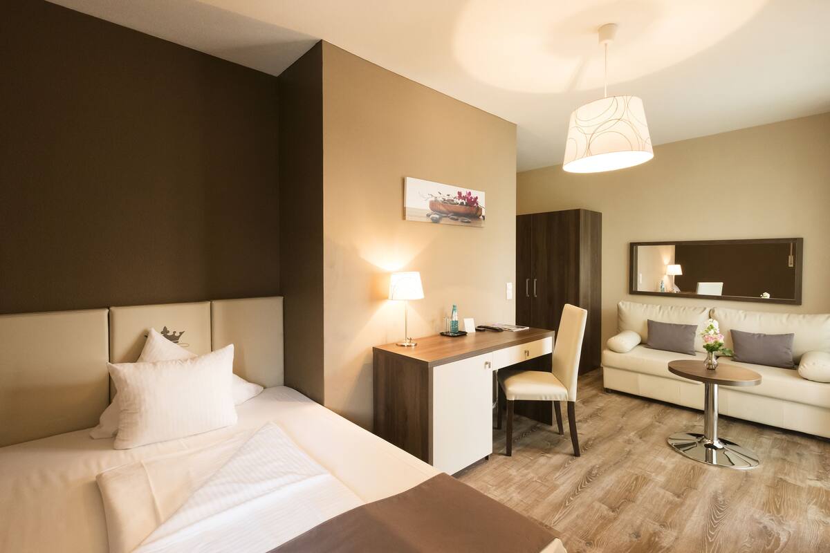 A hotel room with a bed, desk, chair, and a small table with a lamp.