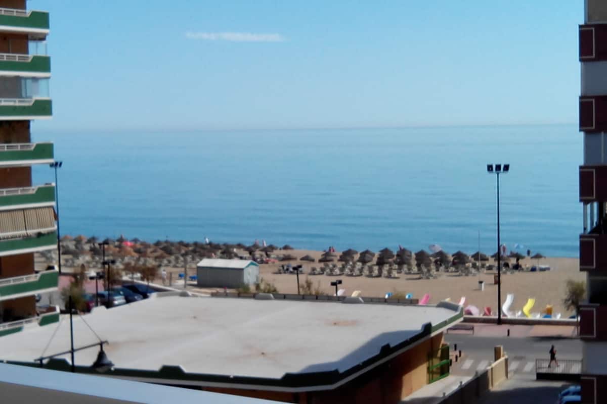 A view of a beach with umbrellas and people, a building with a flat roof, and a clear blue sky.