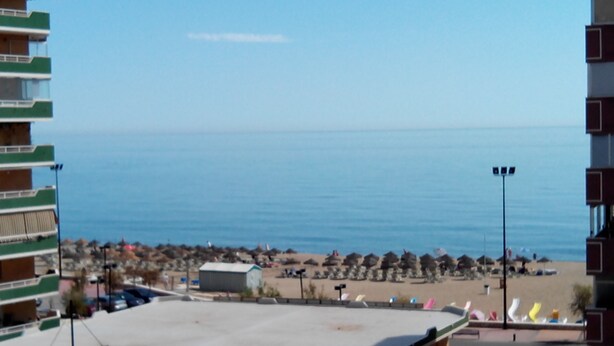 Vista de una playa con sombrillas y gente, un edificio de techo plano y un cielo azul despejado.