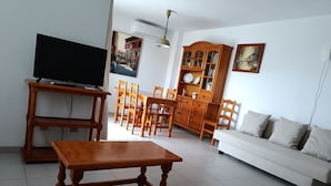 Ein Wohnzimmer mit Fernseher auf Holzständer, ein weißes Sofa und ein Essbereich mit Holzstühlen und einem Schrank.
