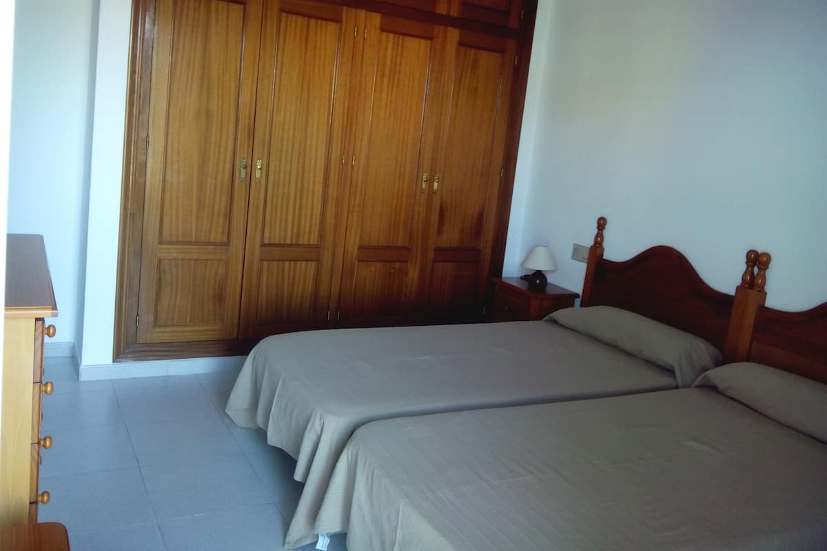 A bedroom with two beds, wooden wardrobe, bedside table with lamp, and a wooden headboard.